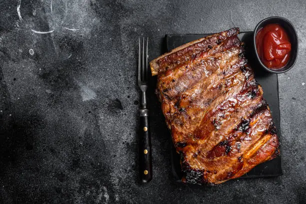 Full rack of BBQ grilled pork spare ribs on a marble board. Black background. Top view. Copy space.