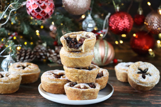 istock Image of white plate containing stack of cooked, homemade mince pies dusted with icing sugar, spruce needles of Christmas tree with red and gold ornaments, blurred white fairy lights background, focus on foreground 1370896746