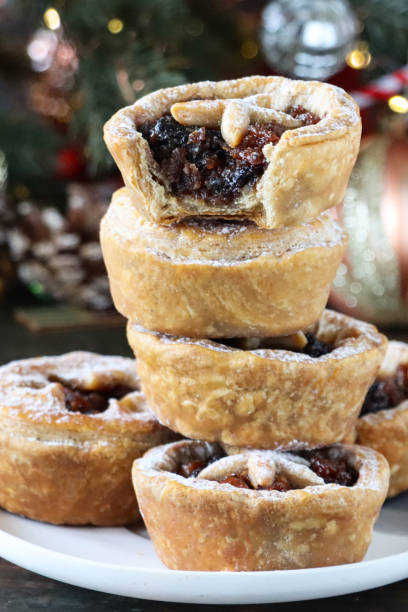 istock Full frame image of white plate containing stack of cooked, homemade mince pies dusted with icing sugar, spruce needles of Christmas tree with red and gold ornaments, blurred white fairy lights background, focus on foreground 1370896747