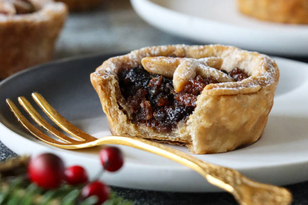 Stock photo showing close-up view of homemade, individual mince pie made with homemade short crust pastry topped with pastry star detail, hiding the sweet mincemeat (mixture of chopped dried fruit, distilled spirits and spices) filling on white plate, with a Spruce Christmas tree decorated with white fairy lights, and red and gold baubles in the background. imagen de fotograma completo de plato blanco que contiene pastel de carne picada casera espolvoreado con azúcar glas con mordida faltante, tenedor de oro, árbol de navidad con adornos rojos y dorados, fondo de luces de hadas blancas, enfoque en primer pl