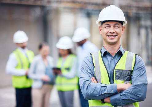 Portrait of man architect at building site with folded arms looking at camera. Confident construction manager in formal clothing wearing white hardhat. Successful mature civil engineer at construction site with copy space.