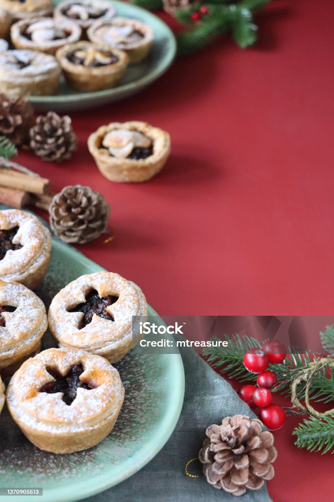 Image homemade mince pies, festive Christmas dessert, crispy pastry filled with sweet mincemeat, topped with star shaped pastry, pine cones, spruce and red berries, red background, focus on foreground, copy space Stock photo showing close-up view of green dishes of individual mince pies made with homemade short crust pastry topped with pastry star detail, hiding the sweet mincemeat filling (mixture of chopped dried fruit, distilled spirits and spices). Mince Pie Stock Photo