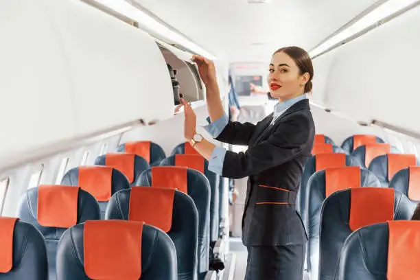 Young stewardess that is in formal black clothes is standing indoors in the plane.