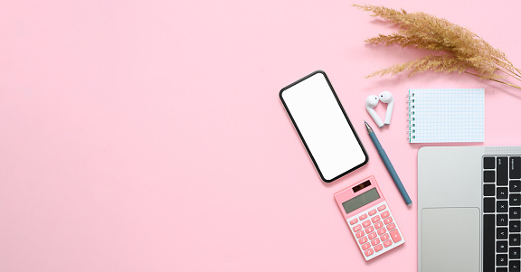 Women's workplace in the office. Smartphone with a blank white display, laptop, notepad and a bunch of dry grass on a pink background. Flat lay, copy space.