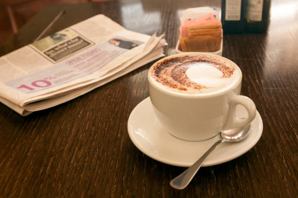 A cup of refreshing morning cappuccino coffee with milk froth and grated cinnamon chocolate on a saucer with a teaspoon, sugar and newspaper on a wooden table