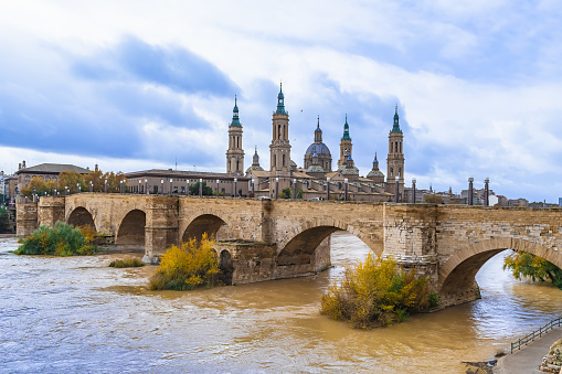 Medieval cityscape of Zaragoza with an ancient stone arched bridge across Ebro river and towers against a blue cloudy sky, Spain. Autumn cityscape with Cathedral-Basilica of Our Lady of the Pillar