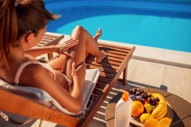 Woman applying sunscreen while sunbathing by the pool Beautiful young woman sunbathing by the swimming pool while on summer vacation, applying sunscreen on her skin as sun UVA and UVB rays protection tanning bed stock pictures, royalty-free photos & images
