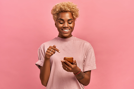 Joyous African American lady with big smile, points at screen of cell phone, rejoices having new app, enjoys online communication, being in high spirit, isolated over pink background. Technology