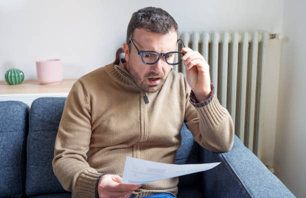 hombre sorprendido leyendo algunas facturas gastos de energía en el sofá sala de estar - energy bill fotografías e imágenes de stock