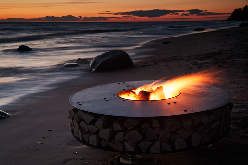 The beautiful metal round a fireplace stands on the sandy coast with a tidal wave at sunset, stones, sand, waves, the landscape, tranquillity, the sun falls, firewood, nobody. High quality photo