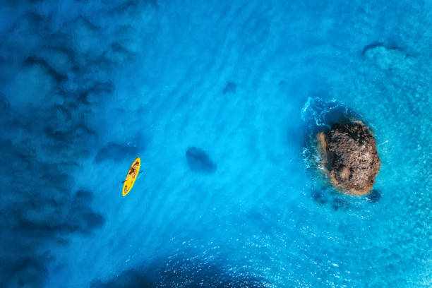 vista aérea del kayak amarillo en el mar azul al atardecer en verano. hombre en canoa flotante en aguas cristalinas, rocas, piedras. isla de lefkada, grecia. paisaje tropical. tabla de sup. viajes activos. vista superior - kayak barco de remos fotografías e imágenes de stock