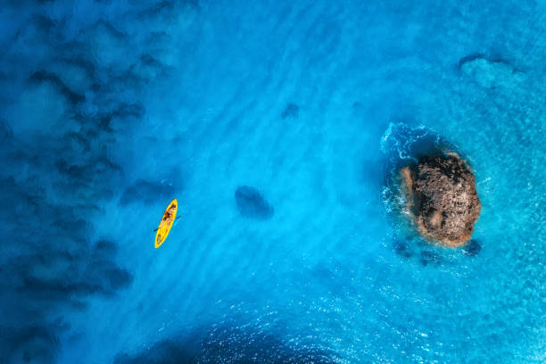 vue aérienne du kayak jaune dans la mer bleue au coucher du soleil en été. homme sur canoë flottant dans une eau azur claire, des rochers, des pierres. île de lefkada, grèce. paysage tropical. sup board. voyage actif. vue de dessus - kayaking kayak sea coastline photos et images de collection