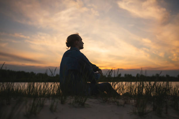 Man silhouette on the background of beautiful summer sunset. Orange sky. Guy is sitting in front of the river. Person sit on the river bank stock photo
