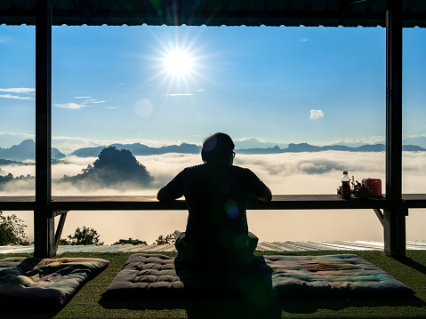 silhouette of man sit at wooden terrace window looking at beautiful view of sea fog on mountains and morning sun at Ban Jabo, noodle restaurant on hill, travel destination in Mae Hong Son, Thailand