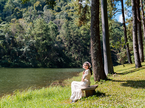 attractive Asian woman in white dress sitting in pine forest riverside at Pang Ung reservoir lake and mountain view in Mae Hong Son, Thailand
