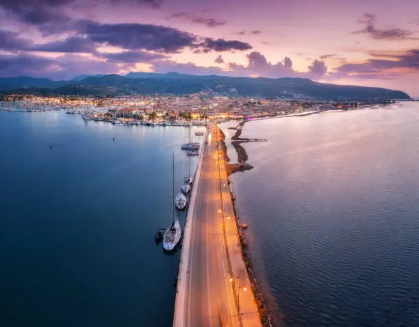 Aerial view of road near sea canal at night in summer in Lefkada island, Greece. Top view of road, blurred cars, boat and yachts, city lights, architecture, mountain and purple sky at sunset. Travel