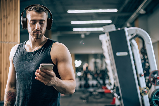 Man is using wireless headphones for music and audio bunk during sports training in fitness club. Men in gym, lifestyle with people indoors. Person with mobile phone for working and fit plan