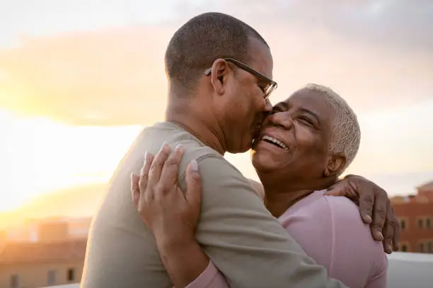 Photo of Happy Latin senior couple having romantic moment embracing on rooftop during sunset time - Elderly people love concept