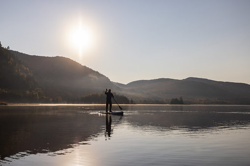 A man is stand-up paddleboarding on Lac Monroe Lake in Mont Tremblant National Park, Quebec, Canada at sunrise. It is a beautiful sunny Autumn day. The mountain is colorful with yellow, red and orange autumn leaves. The man is wearing warm clothing because it is colder in October. There is fog on the lake and the sun is shinning.