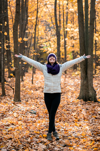 A woman is hiking in the forest in Quebec, Canada. It is a sunny autumn day and colorful leaves are falling from the trees. The woman has her arms raised and smiling. She is very happy and enjoying the moment.
