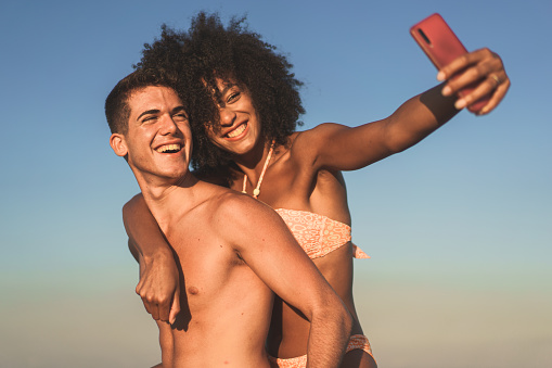 Multicultural couple with boyfriend and girlfriend on vacation taking selfies on the beach - multi-racial young people carefree piggybacking on the beach and taking photos with smartphones in the summerÃ¨