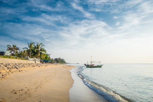 Beautiful view of the turquoise waves with the blue sky and white clouds and fisherman's boat on sandy on vacation in Hua Hin beach, Prachuap Khiri Khan Province, Thailand. Tropical colorful sand from the landscape sea. Summer vacation concept.