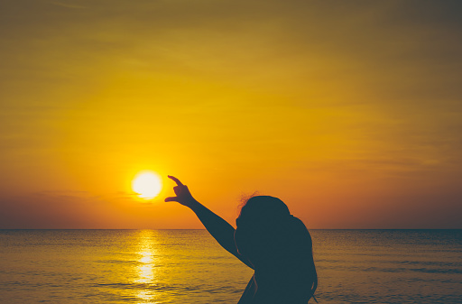 Strong confidence woman raising arms under the sunrise by the landscape sea. Silhouette female enjoying the sunset on the colorful beach. Summer vacation concept