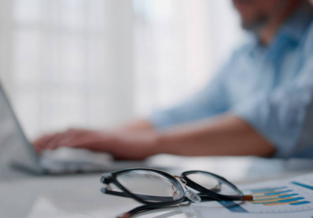 The man is typing on a laptop with a pair of glasses in the foreground. focus in the background