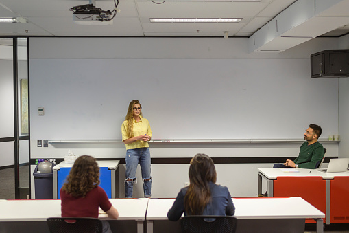 A young woman confidently gives a presentation to a diverse group of university students sitting at desks in a classroom.