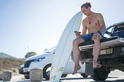 Male surfer sitting on pickup before surf engaging with his mobile