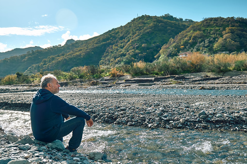 Side view of pensive bearded mature man sitting on riverside of mountain river, contemplating nature. Concept of leisure activities,tourism, lifestyle e nature.