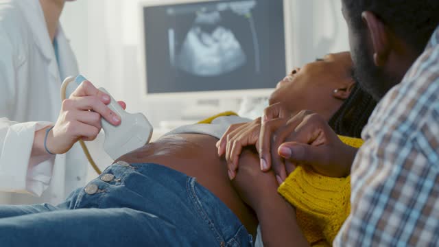 Afro pregnant couple smiling looking at sonogram results on monitor while on checkup at gynecologist