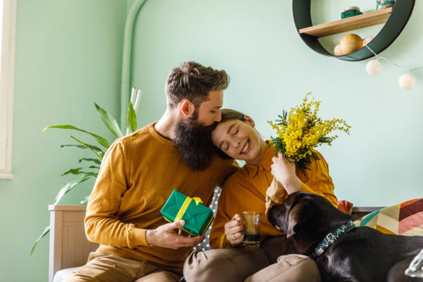 Caring young man kissing his girlfriend on the head after giving her flowers Portrait of affectionate young man embracing and kissing his loving girlfriend after giving her mimosa flowers and a gift box for International women's day. Their brown Labrador is accompanying them. sensitive plant stock pictures, royalty-free photos & images