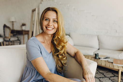 ttractive adult woman sitting on coach in living-room with loft design, having beautiful, clean and combed wavy hair, big blue eyes matching with color of her dress. Natural beauty concept