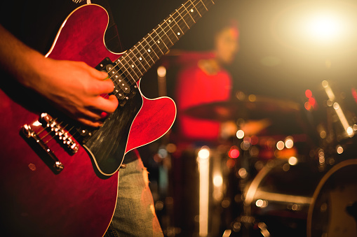 African American vocalist keeps hands on professional microphone stand and sings new composition in sound recording studio. Band plays on musical instruments at background. Music production concept.