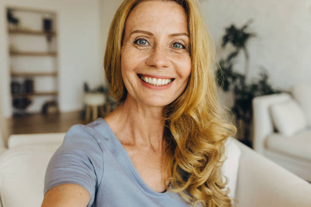 charmante femme âgée soignée faisant un selfie sur son téléphone, regardant l’appareil photo avec un large sourire denté, isolée sur un fond flou de l’intérieur de la maison et de l’étagère. personnes, apparence et concept technologique - toothy smile relaxation white healthcare and medicine photos et images de collection