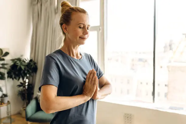 Calmness, wellbeing, peace and spirituality concept. Peaceful smiling senior female in sportswear standing against window in white room making namaste gesture with eyes closed, meditating at home