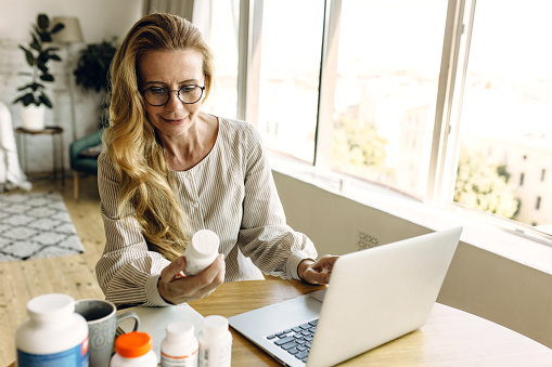 Good-looking elderly female nutrition expert sitting in living-room at table using laptop, holding bottle of vitamins writing article with advices how to select supplements for good healthy skin