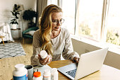 Good-looking freckled ageing woman blogger with long golden hair wearing glasses writing reviews online on new dietary supplements for rejuvenation on her account via social network, using laptop