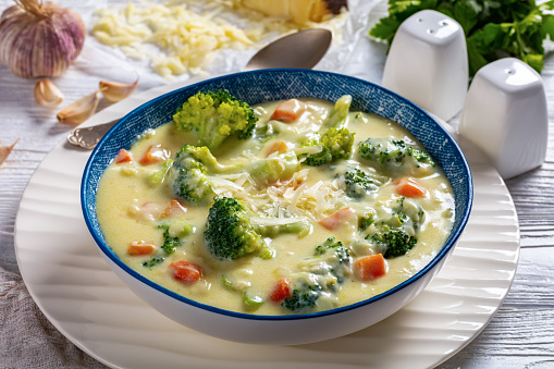 Broccoli cheese soup with chicken broth, cream and carrots in bowl on a white kitchen table with ingredients, horizontal view from above, close-up