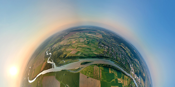 Aerial view from high altitude of little planet earth with small village houses and distant green cultivated agricultural fields with growing crops on bright summer evening.