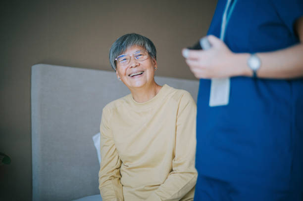 女性看護師の自宅介護者のそばのベッドに座って幸せそうに微笑むアジアの中国人先輩女性 - senior adult women adult tan ストックフォトと画像