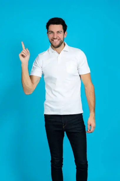 Lifestyle Ideas. Caucasian Handsome Man Posing in White Shirt Looking Straight to Camera And Holding Pointfinger Lifted Over Blue Background.Vertical Image