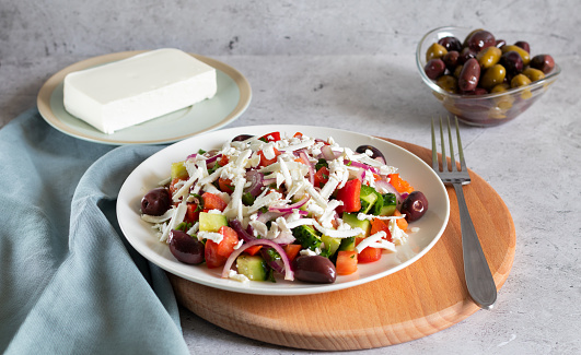 Bulgarian shopska salad with vegetables and cheese on a light concrete background and wooden board with gray textiles and fork. Horizontal, side view.