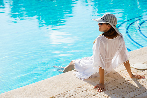 Fashion beautiful woman on summer vacation relaxing at luxury resort spa poolside. Young  fashionable lady wearing sun hat and white kaftan.