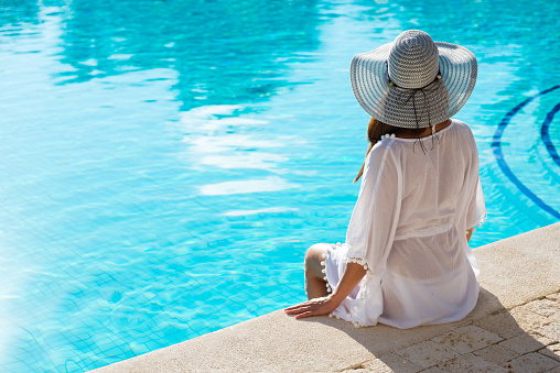 Back view of fashion woman on summer vacation relaxing at luxury resort spa poolside. Young  fashionable lady wearing sun hat and white kaftan.