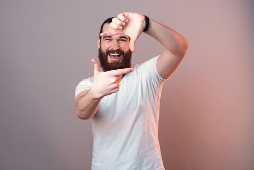 Cheerful bearded man is shaping a camera frame with his fingers. Close up studio shot over grey background.