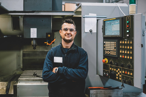 Portrait of happy apprentice engineering worker young man looking at camera posing, working, examining and operating CNC plastic injection molding machinery in factory warehouse after studied manufacturing apprenticeship program certifies XXXL