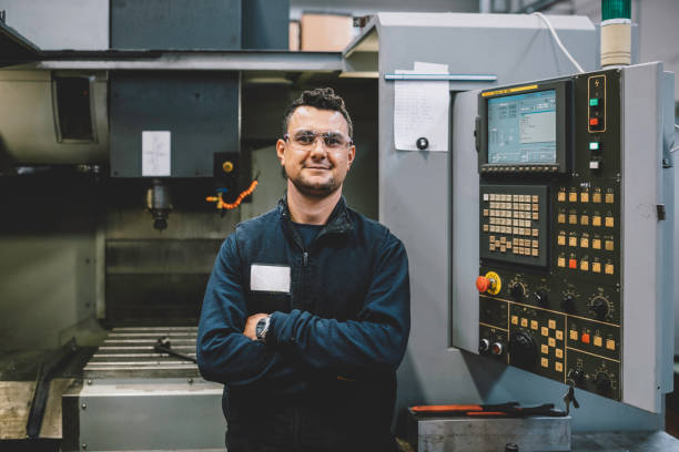 Portrait of smiling male apprentice engineer working with CNC machine in factory Portrait of happy apprentice engineering worker young man looking at camera posing, working, examining and operating CNC plastic injection molding machinery in factory warehouse after studied manufacturing apprenticeship program certifies XXXL manufacturing occupation stock pictures, royalty-free photos & images