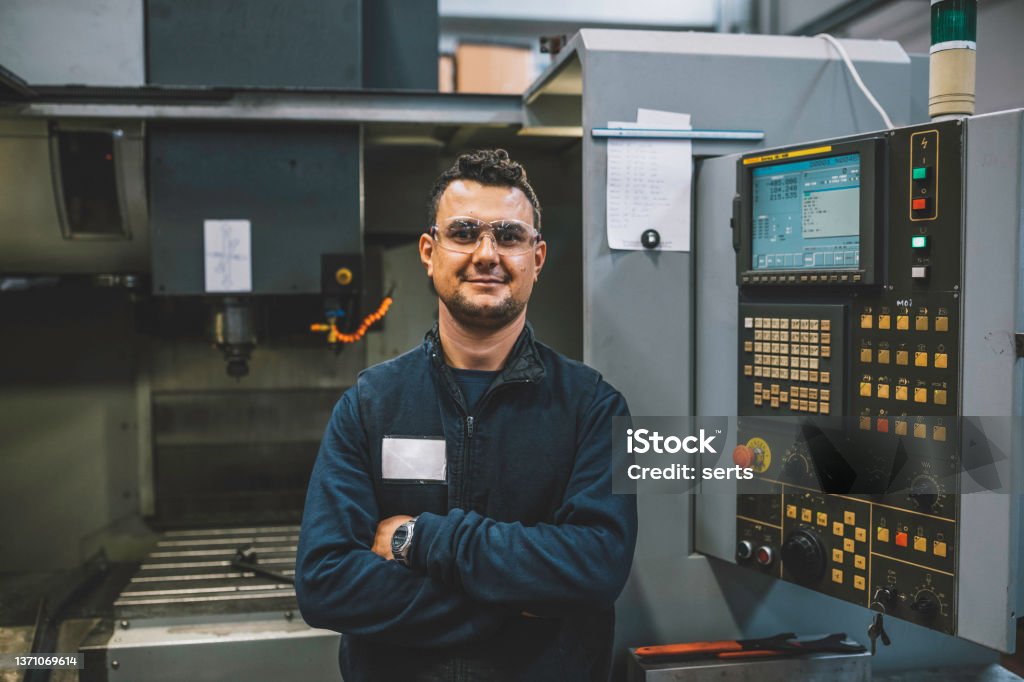 Portrait of smiling male apprentice engineer working with CNC machine in factory Portrait of happy apprentice engineering worker young man looking at camera posing, working, examining and operating CNC plastic injection molding machinery in factory warehouse after studied manufacturing apprenticeship program certifies XXXL CNC Machine Stock Photo